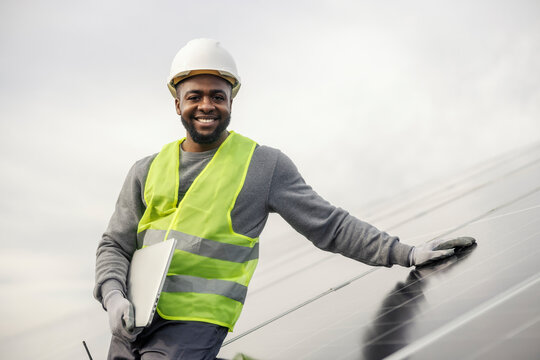 Portrait of smiling diverse serviceman with laptop leaning on solar panel and smiling at camera.