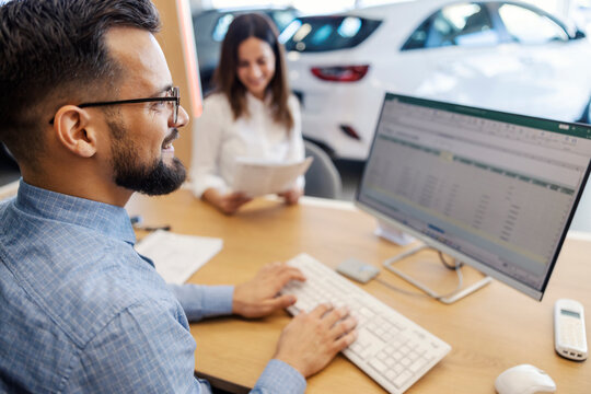 Smiling car dealer sitting at car salon and typing documents on computer while buyer reading contract - Powered by Adobe