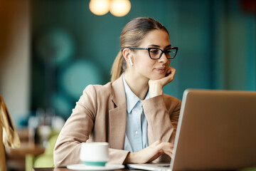 A young woman in suit sitting in coffee shop with headphones and typing searching the internet.