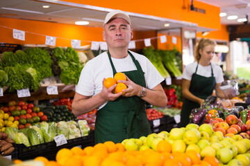 Portrait of a confident salesman standing in the vegetable department of the store near the fruit counter, holding oranges ..in his hands