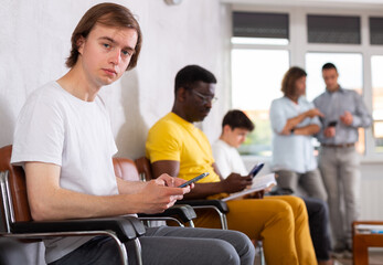 Young male patient sitting on the bench in waiting room of clinic together with other guys