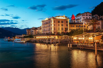 Scenic view of Bellagio at dusk with illuminated buildings along Lungolario Guglielmo Marconi. Calm waters of Lake Como reflect the vibrant evening lights, creating a serene atmosphere