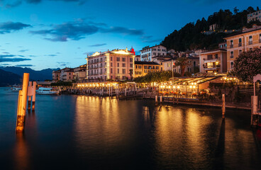 Bellagio's waterfront in Lombardy, Italy, glows with warm evening lights. Charming buildings line the shore, creating a picturesque scene as the water reflects the vibrant ambiance