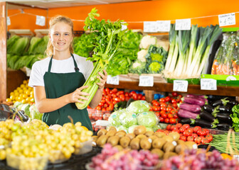Portrait of a positive fifteen-year-old girl who works part-time in a store as a trainee saleswoman, standing in the ..vegetable department, holding celery in her hands
