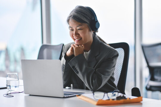 Smiling busy mature professional Asian business woman worker employee wearing suit and headphones looking at laptop computer in office working watching webinar, elearning, having hybrid work meeting.