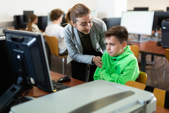 Caring friendly female teacher calming upset tween schoolboy sitting at computer in classroom, helping him complete assignment during lesson