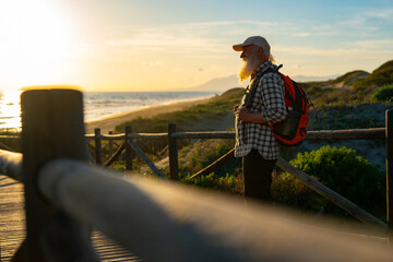 Senior Adventurer with a Red Backpack Admiring the Coastal Sunset on a Scenic Wooden Boardwalk