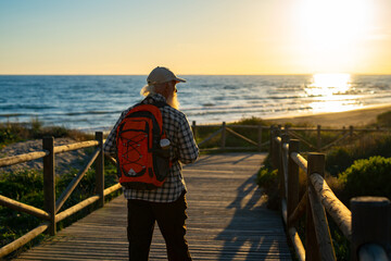 Obraz premium Senior Traveler with a Red Backpack Enjoying a Scenic Sunset Walk on a Wooden Boardwalk by the Ocean, Looking at the Golden Horizon