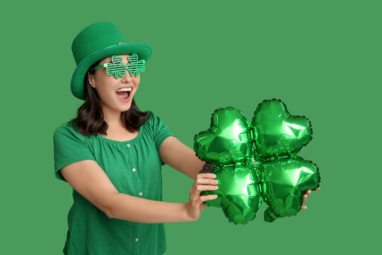Beautiful young woman in leprechaun hat and decorative glasses with air balloon in shape of clover on green background. St. Patrick's Day celebration