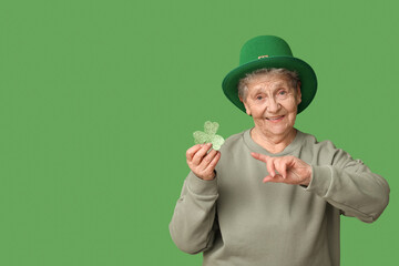 Senior woman in leprechaun hat pointing at clover on green background. St. Patrick's Day celebration