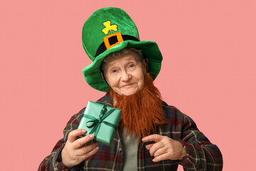 Senior woman in leprechaun hat with red beard holding gift box on pink background. St. Patrick's...