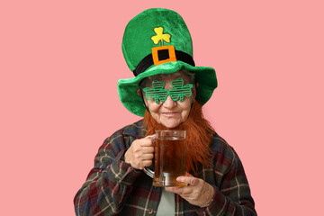 Senior woman in leprechaun hat and decorative glasses with red beard holding glass of beer on pink...