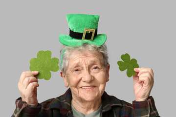 Senior woman in leprechaun hat with clover on grey background. St. Patrick's Day celebration