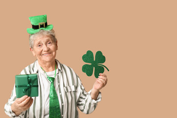 Senior woman in leprechaun hat with clover and gift box on brown background. St. Patrick's Day...