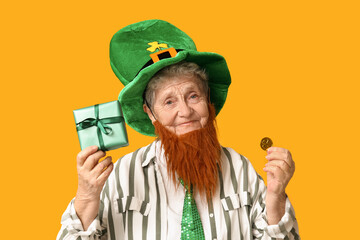 Senior woman in leprechaun hat with red beard holding gift box and coin on yellow background. St....