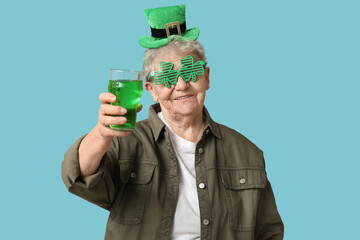 Senior woman in leprechaun hat and decorative glasses with glass of beer on blue background. St....