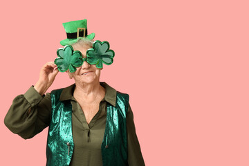 Senior woman in leprechaun hat and decorative glasses in shape of clover on pink background. St....