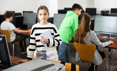 Portrait of diligent interested teen schoolgirl ready for lesson in computer class, standing near table with workbook in hands..