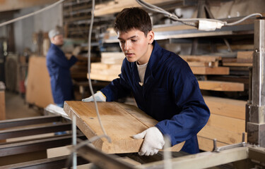Young male carpenter putting wooden board on cutting machine in wood workshop