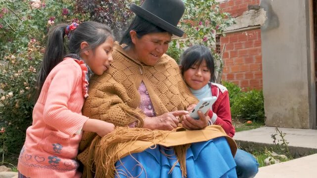 aymara cholita grandmother in pollera looking at her cell phone with her two granddaughters in the backyard of her house in the city of la paz bolivia - communication concept