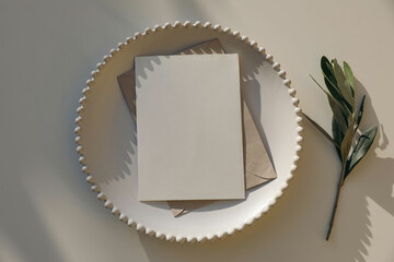 Blank vertical greeting card, invitation mockup, craft envelope in sunlight on white plate. Beige...