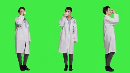 Woman physician drinking a cup of coffee against greenscreen backdrop, taking a break from medical work. Specialist serving caffeine refreshment in studio, wearing a white coat. Camera A.