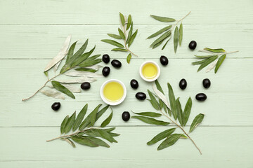 Bowls of oil, tasty black olives and leaves on green wooden background