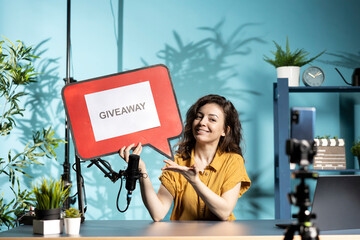 Cheerful vlogger advertising giveaway for viewers with cardboard sign, filming with smartphone on...