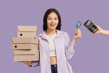 Young Asian woman with shoe boxes and credit card paying via terminal on lilac background