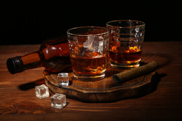 Glasses of rum with ice cubes and cigar on wooden table against dark background
