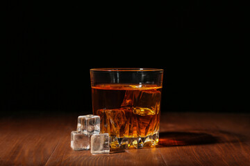 Glass of rum with ice cubes on wooden table against black background