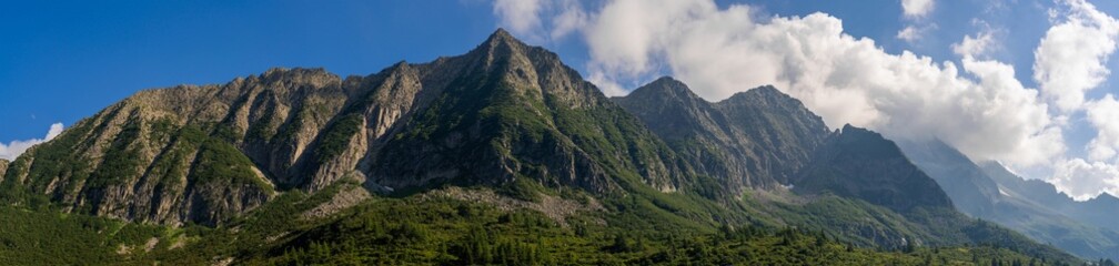 Panoramic shot of stunning mountain range with rugged peaks and lush greenery, blue sky with clouds, forest, alpine landscape. Tonale Pass, Italy. Summer vacation. Dolomite Alps. Ecology concept