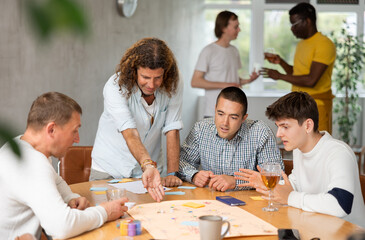 Group of enthusiastic men playing board game sitting around large table while active player is...