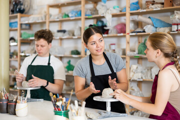Young woman teacher shows students teenage girl and teenage boy how to sculpt ceramic product from raw clay in workshop