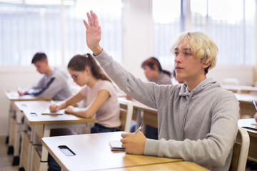 Fototapeta premium Portrait of smart teenage schoolboy raising hand to answer during lesson in college