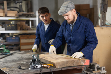 Carpenter measures the length of board with a tape measure. Foreman prompts him