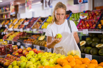 Focused fifteen-year-old girl who came to the store for shopping, chooses ripe apples, standing at the counter