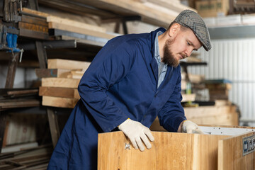 Young male carpenter assembles wooden cabinet from parts in wood workshop