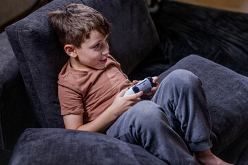 Fototapeta premium Boy is sitting on sofa at home and playing video games with his joypad.