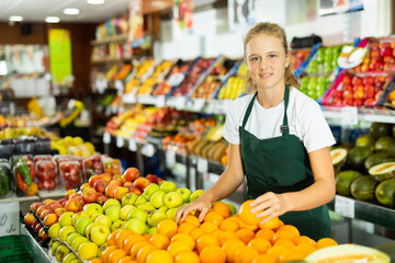 Hardworking fifteen-year-old girl who works part-time in a store as a trainee saleswoman puts oranges on the counter