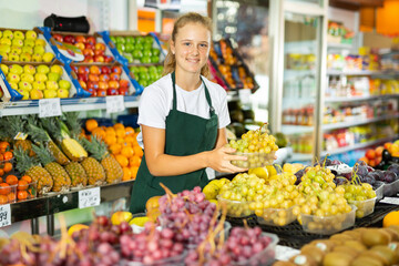 Fifteen-year-old girl who works part-time in a store as a trainee saleswoman puts ripe grapes on the counter, laid out..in boxes