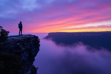 Fototapeta premium Silhouette of hiker on clifftop overlooking a misty valley at sunset.