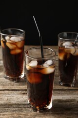 Refreshing cola with ice cubes and drinking straws in glasses on wooden table against black background, closeup