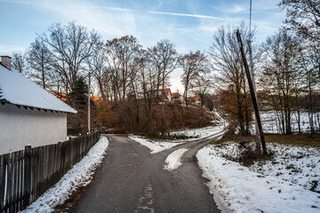 A rural street in a Serbian village is dusted with winter snow, revealing modest homes and leafless...