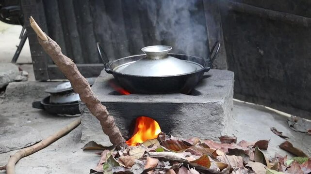 Curry cooking on a traditional clay stove fueled by dry leaves and cow dung sticks in a Bangladeshi village. Covered with a silver lid, steam rises as it cooks, creating a yellow flame and smoke.