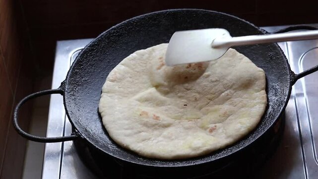 Cooking flatbread or paratha on an iron tawa, gently pressing and turning with a steel spatula on the kitchen stove for a delicious morning breakfast.