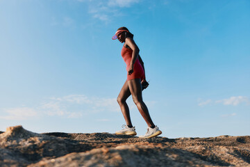 Woman hiking on rocky terrain under a clear blue sky, wearing athletic clothes and a visor,...