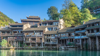 Naklejka premium Old wooden houses of the ancient Fenghuang city. Dilapidated buildings on stilts on the riverbank. Curved tiled roofs against the blue sky. Green vegetation. China. Tuojiang River