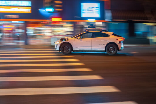 LOS ANGELES, CA - February 12, 2025: Waymo driverless self driving taxi cab rushing through city traffic, crossing illuminated street intersection at night  in West Hollywood, California.