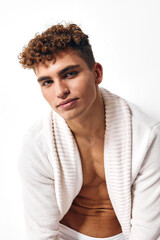 Confident young man with curly hair wearing a white bathrobe, showcasing a relaxed expression against a clean white background. Spa and self-care concept.